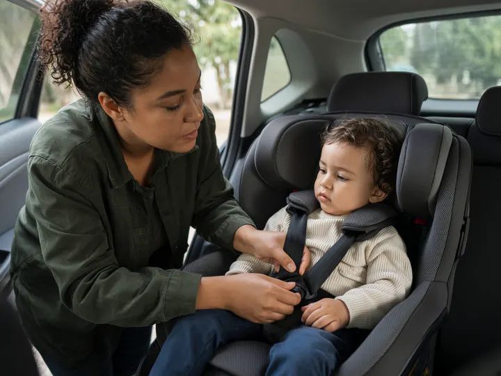 An adult in a dark green shirt leans into a car and buckles a toddler in a cream knit sweater into a harnessed car seat.